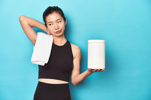 Jovem desportista asiática com uma garrafa de proteína no ginásio a tocar a parte de trás da cabeça a pensar.