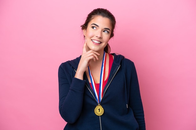 Jovem desportista brasileira com medalhas isoladas em fundo rosa pensando em uma ideia enquanto olha para cima