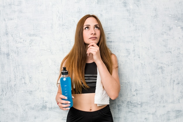 Jovem desportista caucasiana segurando uma bebida isotônica, olhando de soslaio com expressão duvidosa e cética.