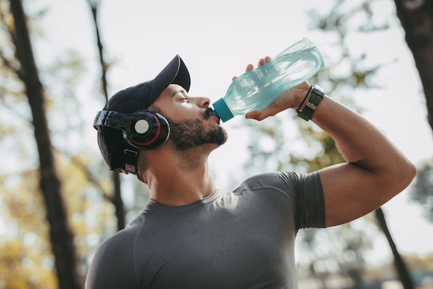 Foto jovem desportista com água potável de fones de ouvido após o treino no parque.