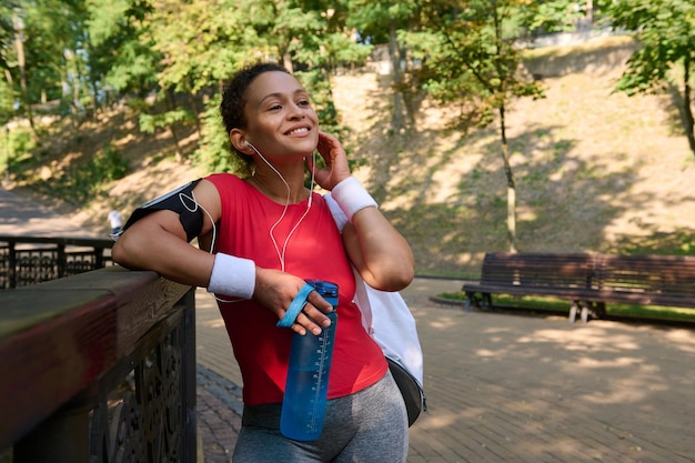 Jovem desportista em activewear sorri lindo sorriso dentuço segurando uma garrafa com água e colocando fones de ouvido relaxando após treino funcional e cardio de corrida matinal no parque da cidade