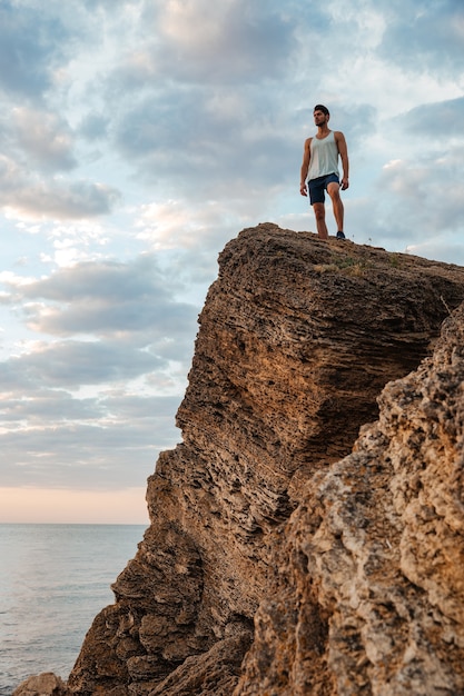 Jovem desportista em pé na rocha da montanha à beira-mar e olhando o pôr do sol