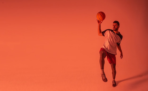 Jovem desportista europeu joga basquete. Jogador de basquete barbudo bonito usa roupas esportivas. Isolado em fundo laranja. Sessão de estúdio. Copie o espaço