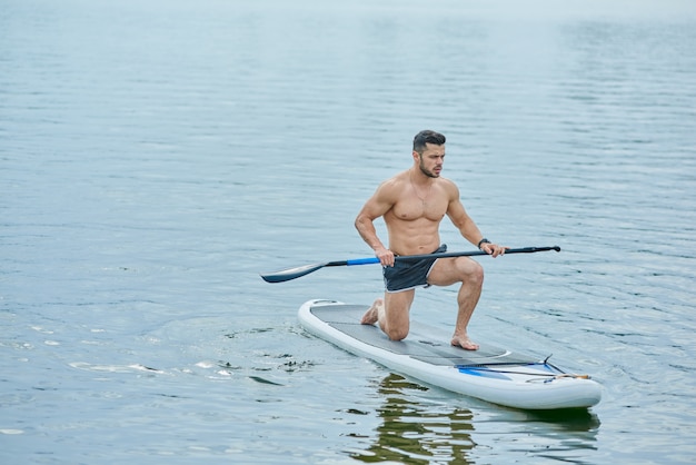 Jovem desportista mantendo longo remo, de pé sobre um joelho, nadando a bordo de sup no lago da cidade.