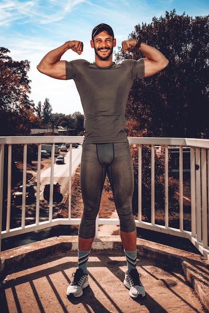 Foto jovem desportista sorridente urbana apontando seu músculo após o treino na ponte e olhando para a câmera.