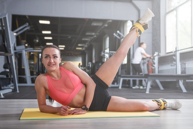 Jovem desportiva alegre desfrutando de se exercitar na academia, levantando as pernas com pesos nos tornozelos