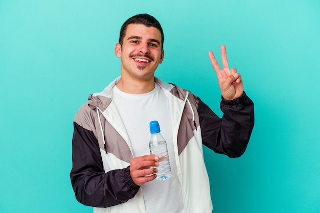 Jovem desportivo bebendo água isolada na parede azul, alegre e despreocupado, mostrando um símbolo da paz com os dedos