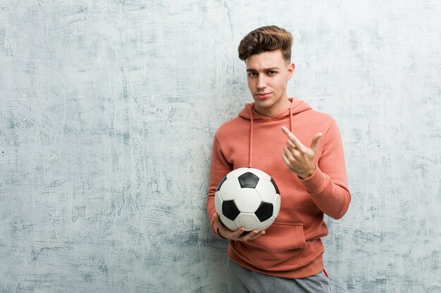 Jovem desportivo segurando uma bola de futebol, apontando com o dedo para você, como se convidando se aproximar.