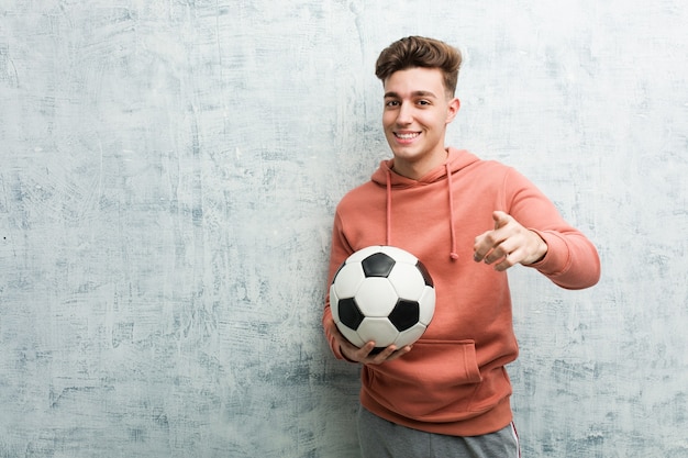 Jovem desportivo segurando uma bola de futebol, sorrisos alegres, apontando para a frente.