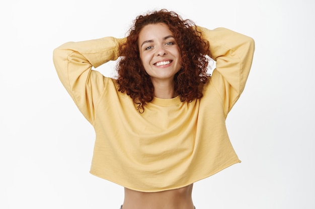 Foto jovem despreocupada com cabelo ruivo encaracolado, olhando relaxado e sorrindo feliz para a câmera, mãos atrás da cabeça, mentindo e descansando, posando contra fundo branco