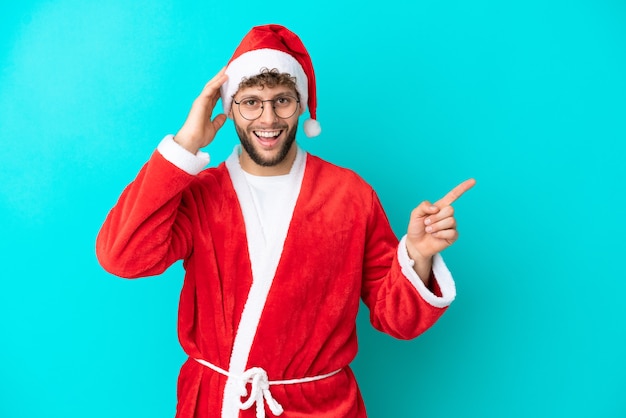 Jovem disfarçado de Papai Noel isolado em um fundo azul surpreso e apontando o dedo para o lado