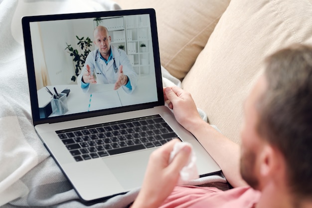 Jovem doente com laptop e lenço amassado sentado no sofá debaixo do cobertor enquanto descansa e consulta um médico online