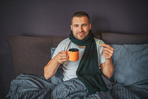 Jovem doente, mas positivo, senta-se na cama no quarto e parece camer. Ele segura um copo de laranja em uma mão e um pedaço de limão em outra. Cara sorri. Ele parece feliz.