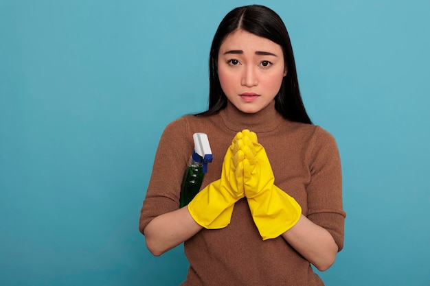 Jovem dona de casa asiática assustada juntou as mãos usando luvas amarelas e um pulverizador de detergente sob o braço isolado contra um fundo azul, trabalhadora dona de casa, conceito de limpeza em casa,