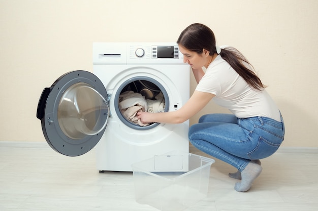 Jovem dona de casa coloca roupas sujas na máquina de lavar moderna de uma caixa de plástico perto da parede bege