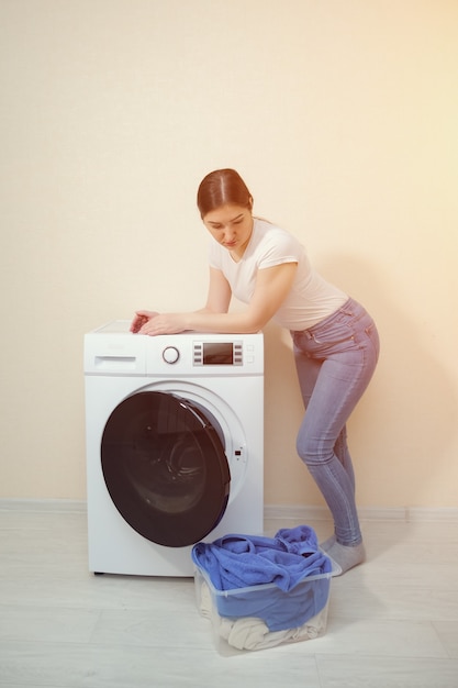 Jovem dona de casa encostada na moderna máquina de lavar, olhando para uma caixa com roupa suja perto da parede bege de casa