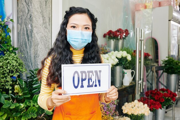 Jovem dona de floricultura usando máscara médica na entrada com placa aberta