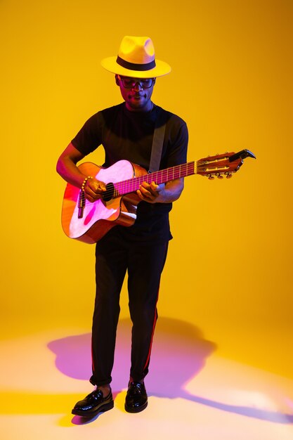 Jovem e alegre músico afro-americano, tocando violão e cantando no fundo gradiente do estúdio laranja-amarelo em luz de néon. Conceito de música, hobby, festival. Retrato colorido do artista moderno.