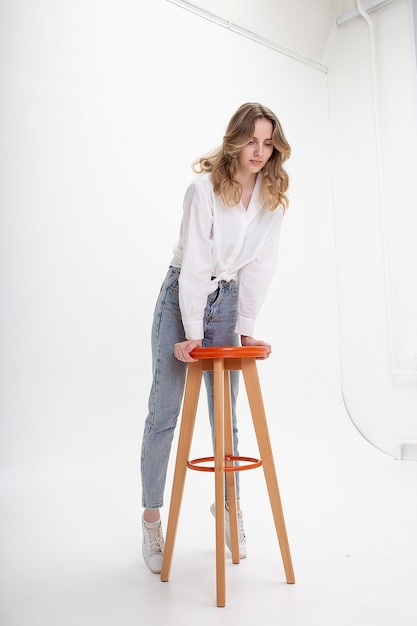 jovem e atraente mulher sorridente com cabelo comprido na camisa, jeans no fundo branco do estúdio pulando no banquinho