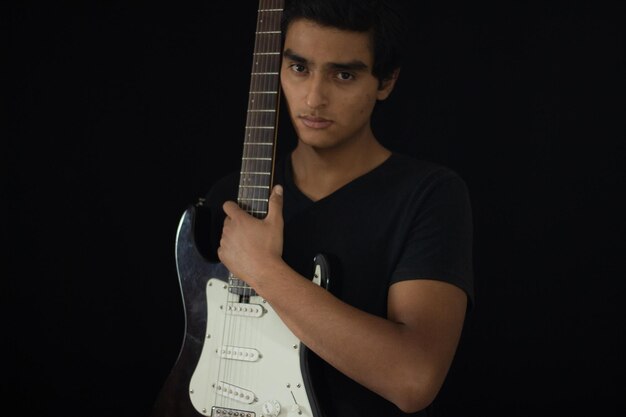 Jovem e atraente músico latino posando dentro de um estúdio e abraçando sua guitarra elétrica e olhando para a câmera.