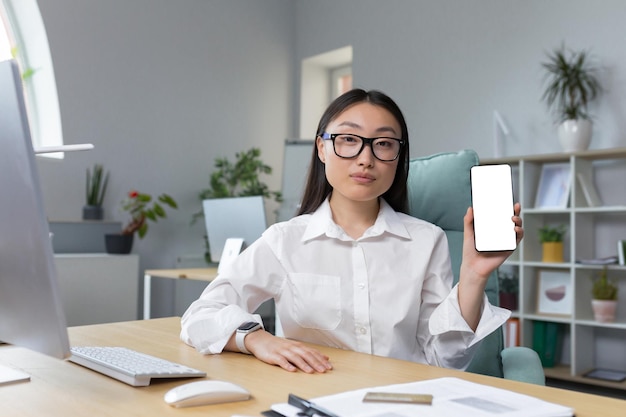 Jovem e bela empresária asiática trabalhando no escritório olhando para a câmera oferecendo telefone aos clientes