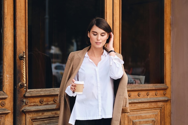 Jovem e bela empresária elegante com café para ir sonhadoramente ao ar livre