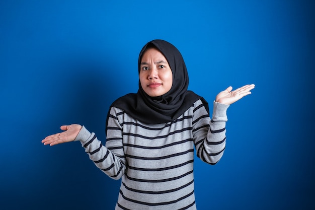 Jovem e bela estudante muçulmana asiática encolhe os braços, faz um gesto de não sei, não posso evitar nada contra o fundo azul