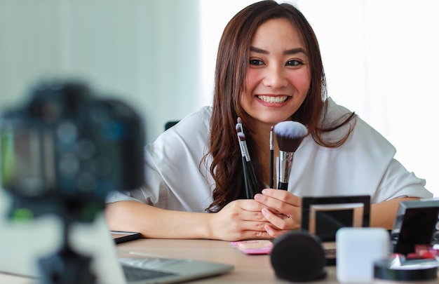 Jovem e bela garota asiática mostra vários pincéis faciais para a câmera com um sorriso e uma alegria durante a transmissão da gravação do vídeo sobre o conteúdo e a revisão de cosméticos. Venda online e conceito de marketing.