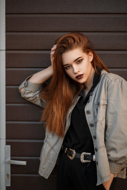Jovem e bela modelo mulher com uma jaqueta elegante e polo preta posa perto do portão na rua