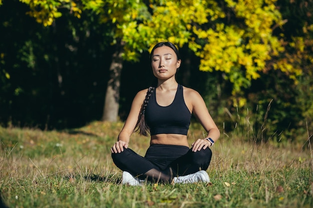 Jovem e bela mulher asiática realiza exercícios de ioga fitness, sentado em posição de lótus na grama e meditando no parque