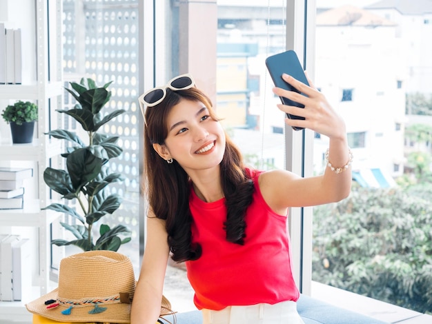 Jovem e bela mulher asiática sorridente segurando o telefone celular inteligente e fazer selfie pela câmera Mulher asiática feliz usando óculos escuros e chapéu de praia tirar uma foto animada viagem de férias de verão