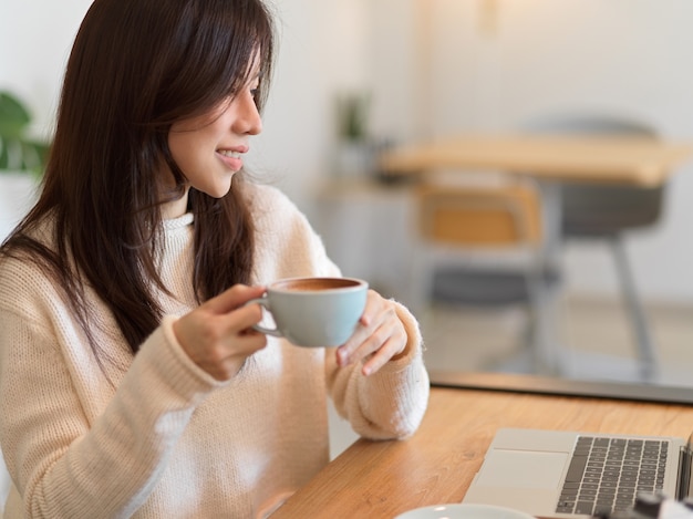 Jovem e bela mulher bebendo café da manhã enquanto trabalhava no computador laptop no café bar.