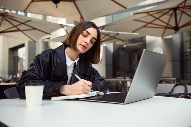 Jovem e bela mulher caucasiana, redator escrevendo no bloco de notas, usando laptop sentado à mesa com um copo de papel de café. Conceito de negócios online, trabalho remoto freelance, aprendizagem à distância pela Internet