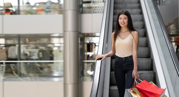 Jovem e bela mulher coreana desce a escada rolante no shopping. sacolas de compras brilhantes. lugar para texto. copie o espaço. baner