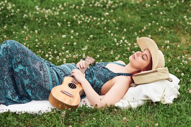 Jovem e bela mulher está descansando deitada na grama em um dia de verão, segurando um ukulele.