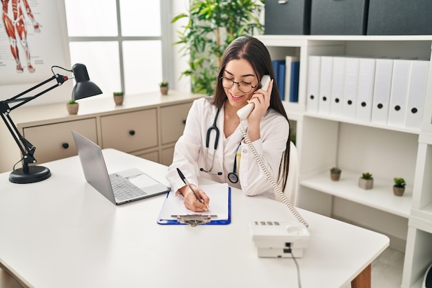 Jovem e bela mulher hispânica, médica, a falar ao telefone, a escrever um documento na clínica.