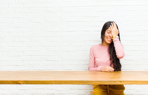 Jovem e bela mulher latina rindo e batendo na testa como dizer dããããããão! Eu esqueci ou isso foi um erro estúpido sentado na frente de uma mesa