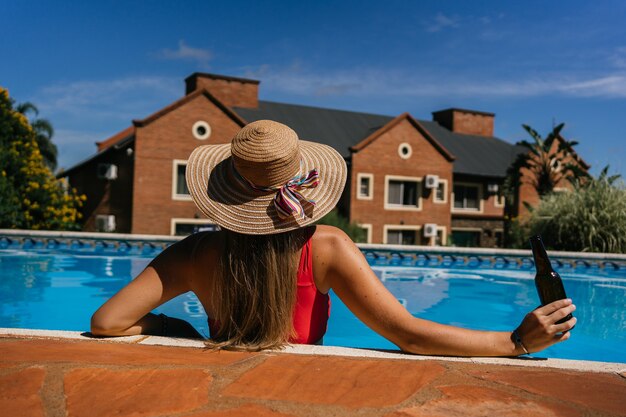 Foto jovem e bela mulher relaxando à beira da piscina com uma garrafa de cerveja.