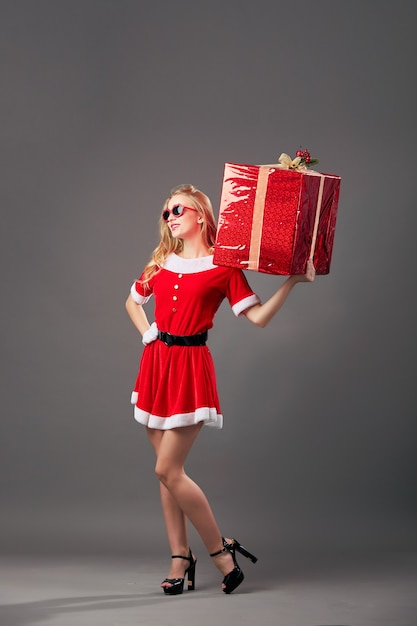 Jovem e bela Senhora Santa Claus em óculos de sol vestida com o manto vermelho, luvas brancas e salto alto detém o enorme presente de Natal no fundo cinza. .