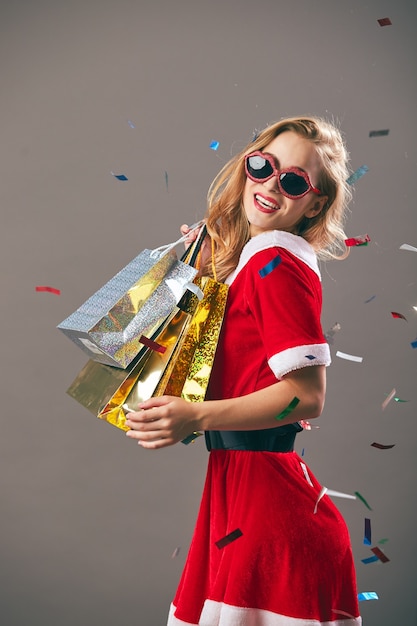 Jovem e bela Sra. Santa Claus em óculos de sol vestida com o manto vermelho e luvas brancas detém os sacos com presentes e sorrisos no fundo cinza com confete. .