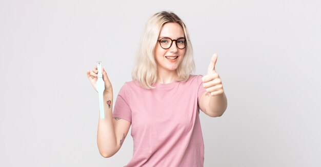 Jovem e bonita albina se sentindo orgulhosa, sorrindo positivamente com o polegar para cima segurando um relógio