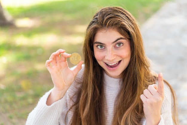 Jovem e bonita mulher caucasiana segurando um Bitcoin ao ar livre apontando uma grande ideia