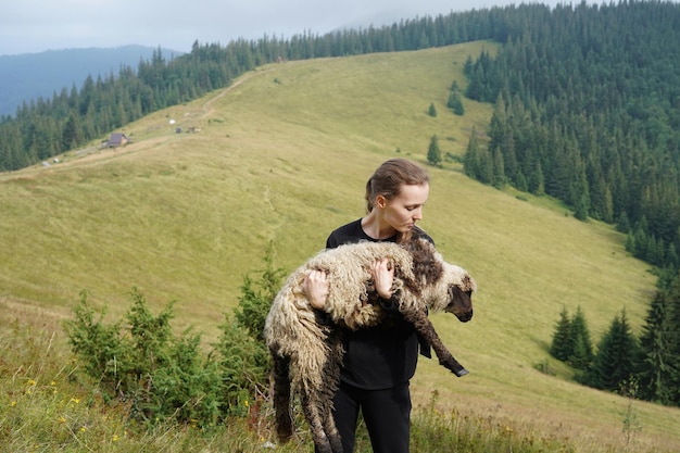 Jovem e bonita pastora segurando uma ovelha no meio de uma paisagem montanhosa o conceito de cuidar de animais, agricultura responsável e amor pela natureza
