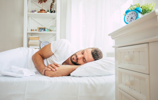 Jovem e bonito barbudo dormindo em uma grande cama branca em um quarto confortável