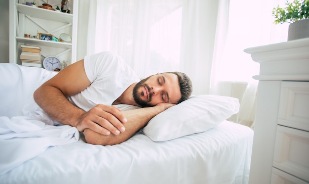 Jovem e bonito barbudo dormindo em uma grande cama branca em um quarto confortável