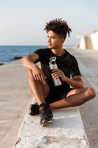 Jovem e bonito desportista sentado ao ar livre na praia a beber água