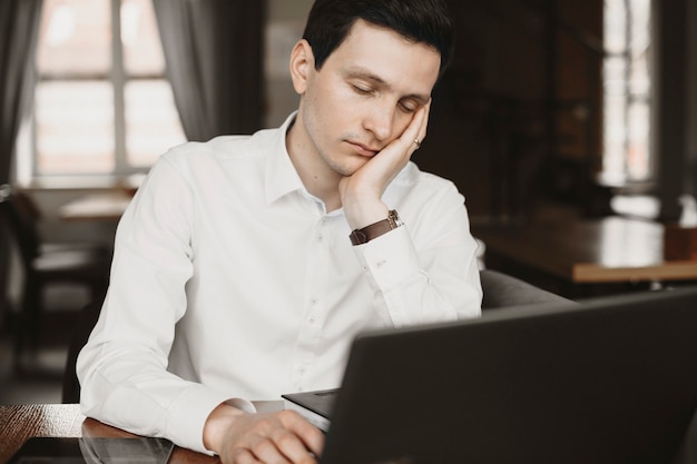 Jovem e bonito freelancer masculino escorregando inclinado na mão enquanto trabalhava em um café em seu laptop.