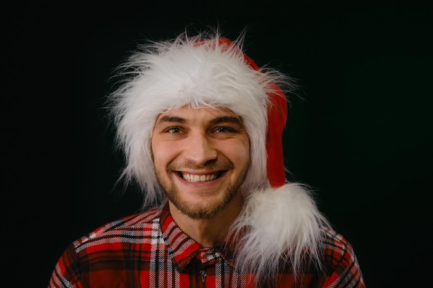 Jovem e bonito homem caucasiano com chapéu vermelho fofo de Papai Noel ri sobre o fundo escuro do estúdio