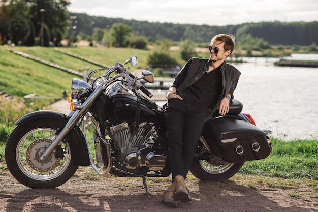 Jovem e bonito motociclista está sentado em sua moto ao ar livre no fundo do lago
