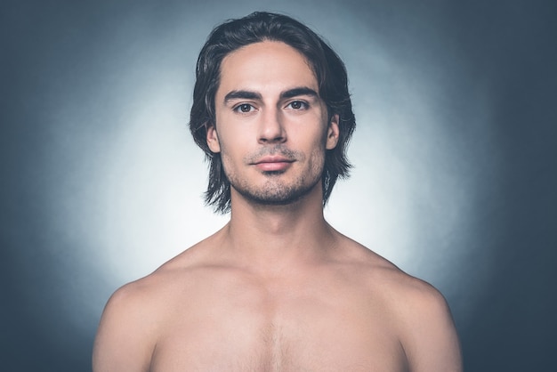 Jovem e bonito. Retrato de jovem sem camisa, olhando para a câmera em pé contra um fundo cinza